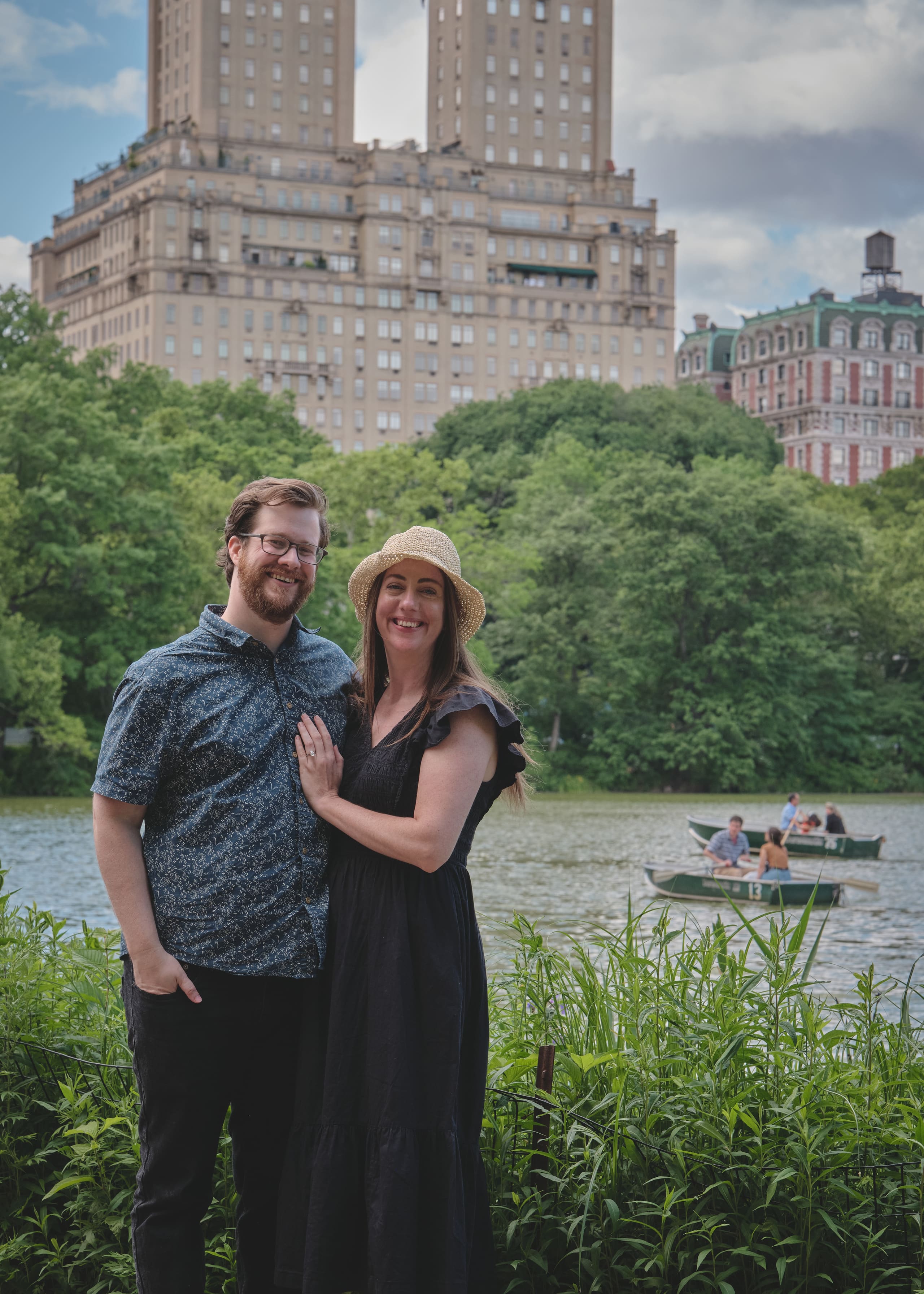 Newly engaged by the lake in Central Park, rowboats and city towers behind them