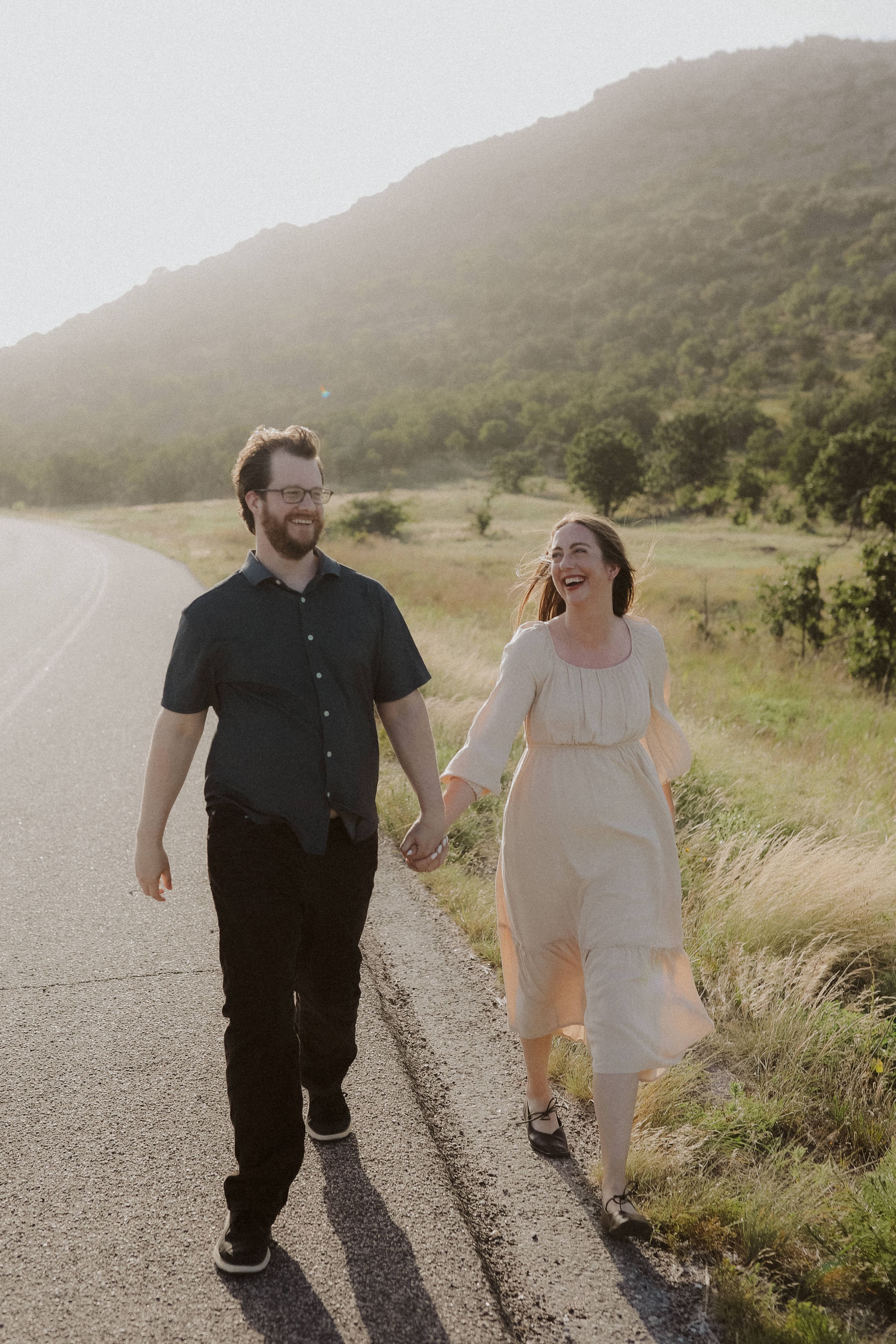 The couple laughing and holding hands, walking down a country road toward the hills
