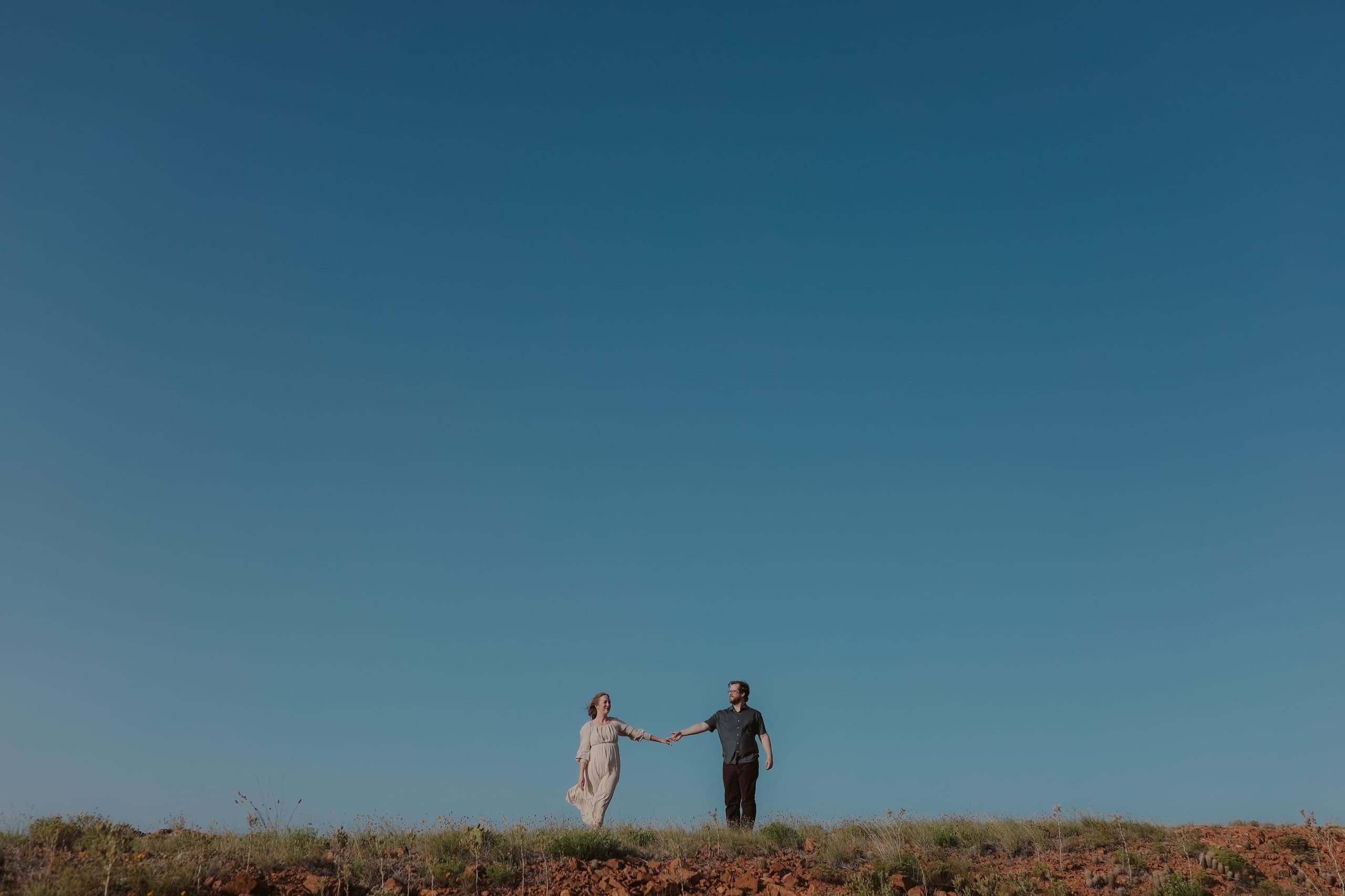 The couple holding hands at arm’s length on a prairie ridge beneath a vast evening sky