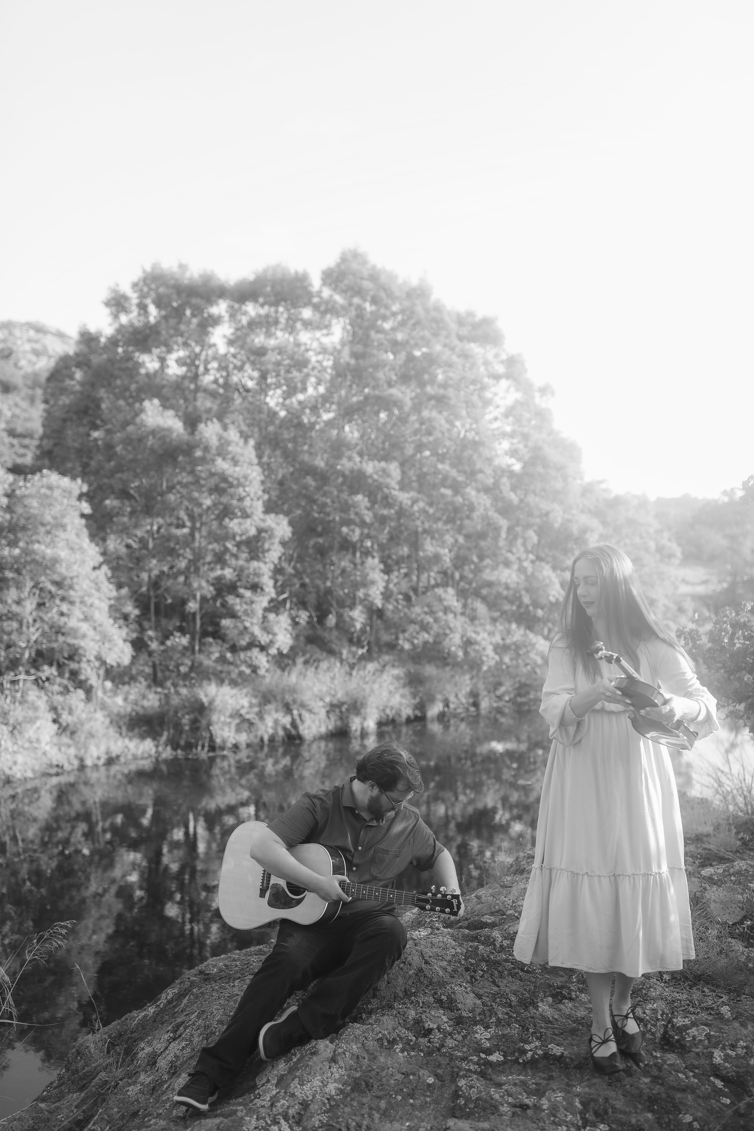 He picks the guitar while she stands with her violin by the water