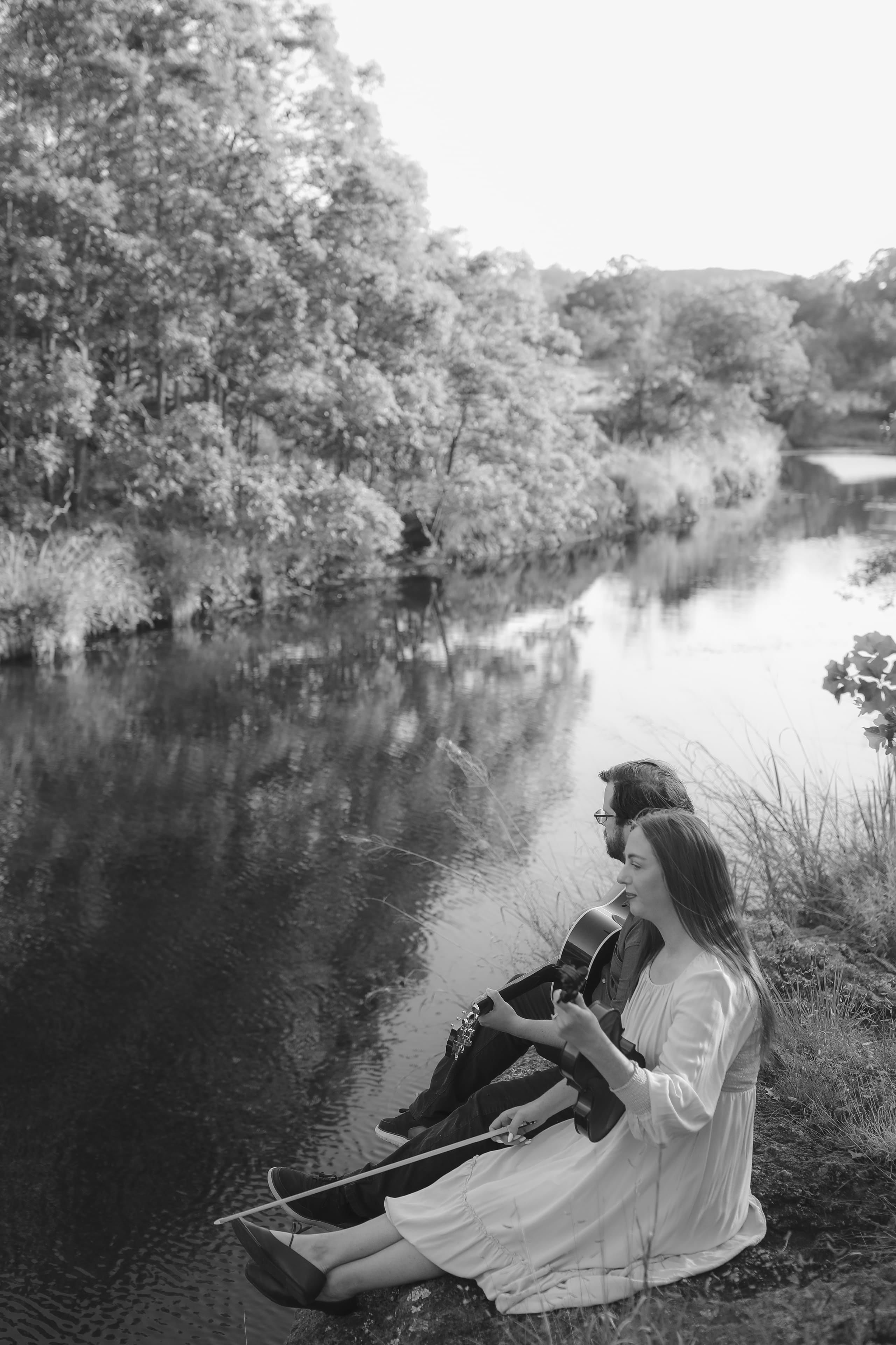 Seated on the riverbank in black and white, playing as the water mirrors the trees