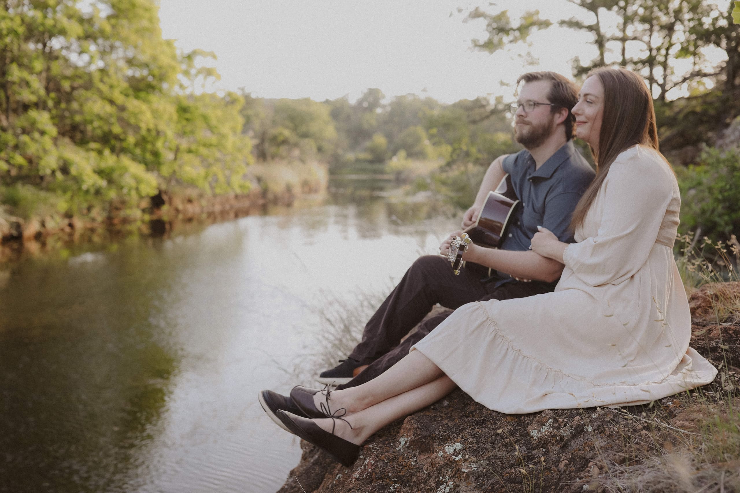 She holds his arm while he plays guitar on the rocks above the river