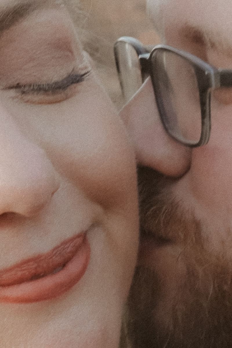An extreme close-up — her quiet smile against his cheek, glasses catching the light