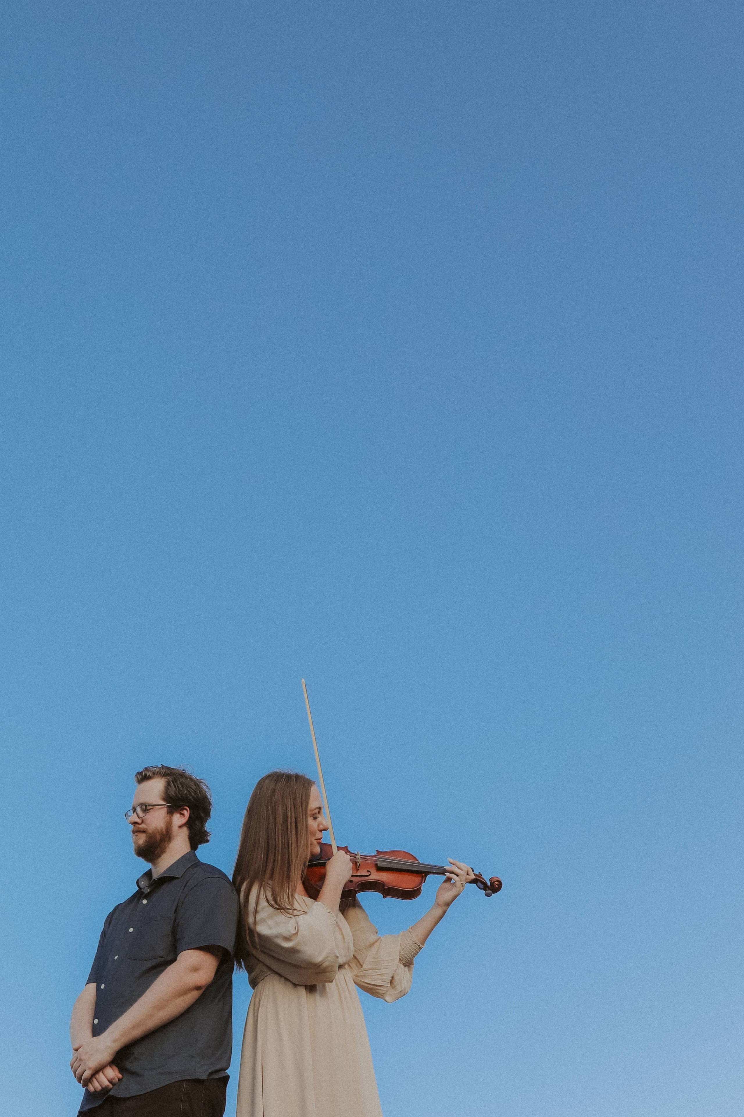 She plays violin at his back beneath a wide clear sky