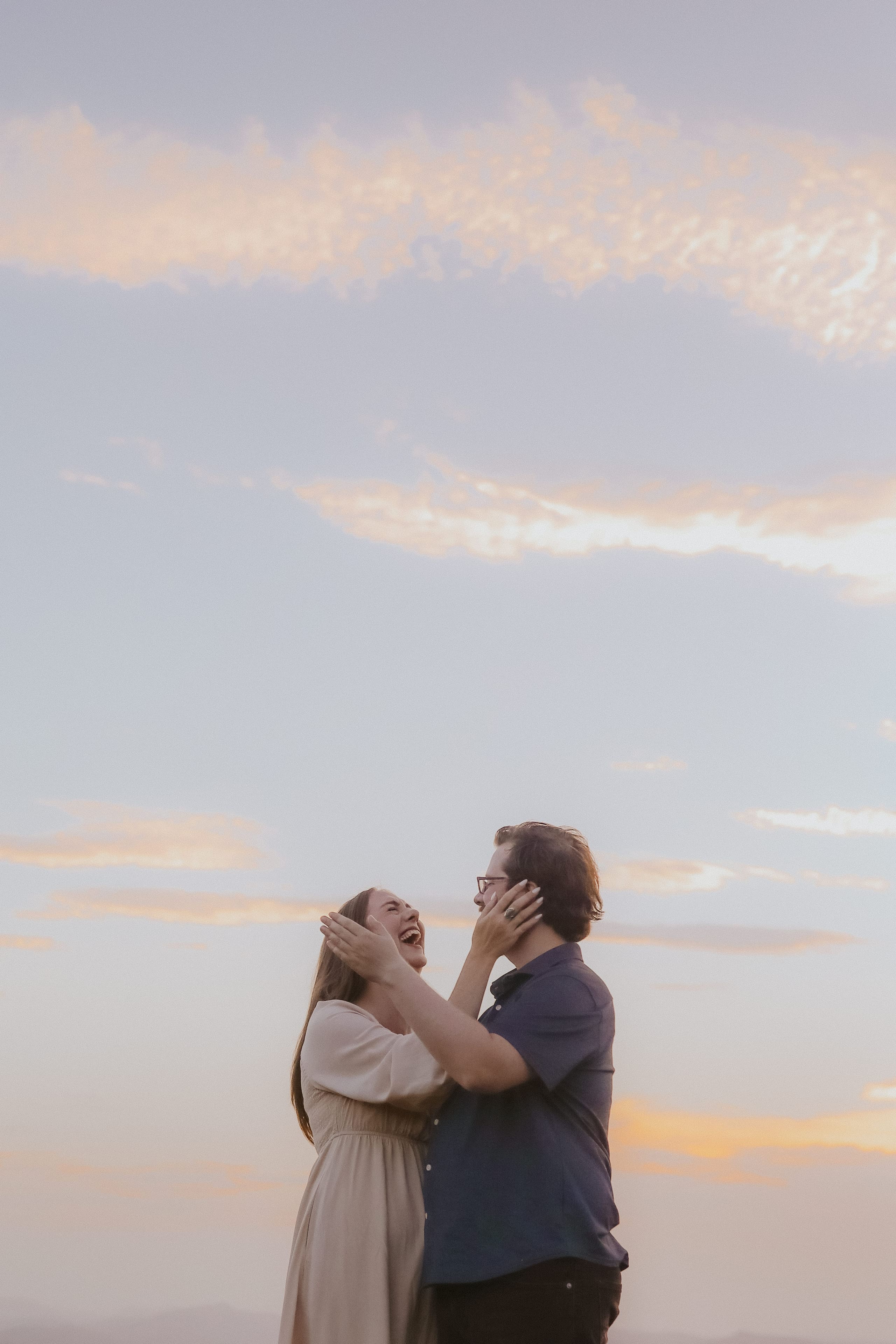 Laughing with her hands on his cheeks under a pastel evening sky