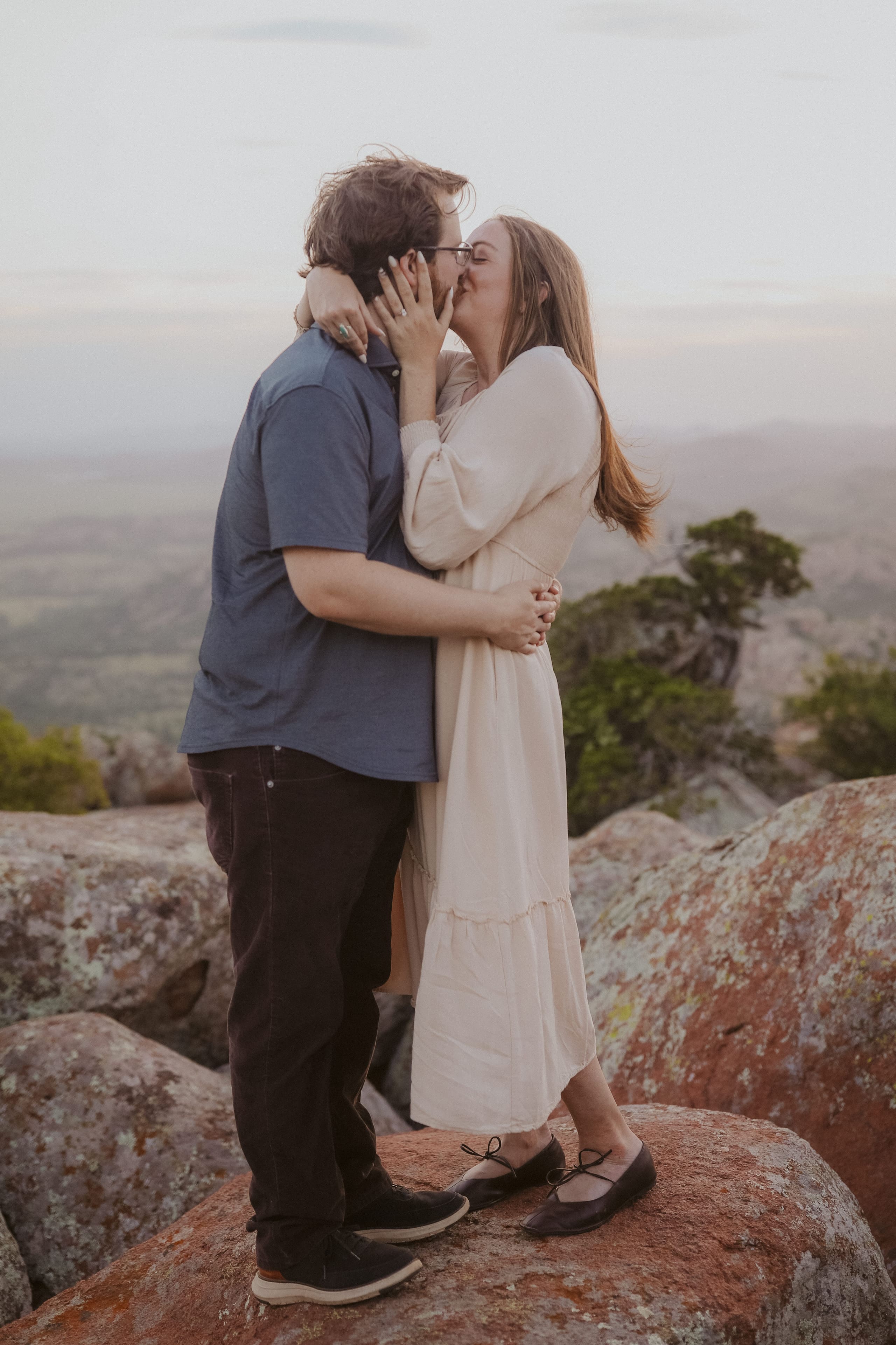 Her hand on his cheek mid-kiss atop the lichen-speckled rocks