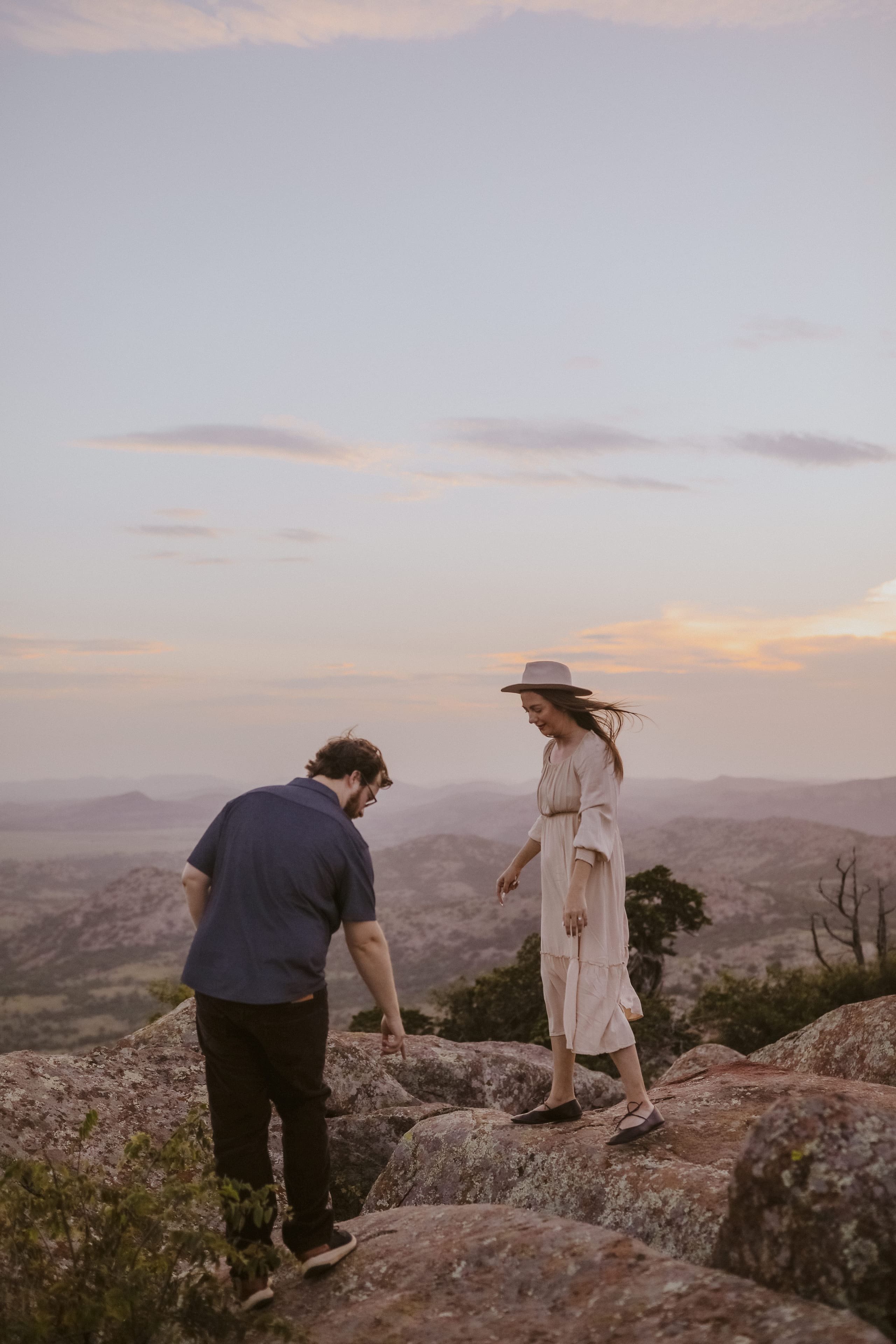 He steps down the rocks ahead as she follows in her hat