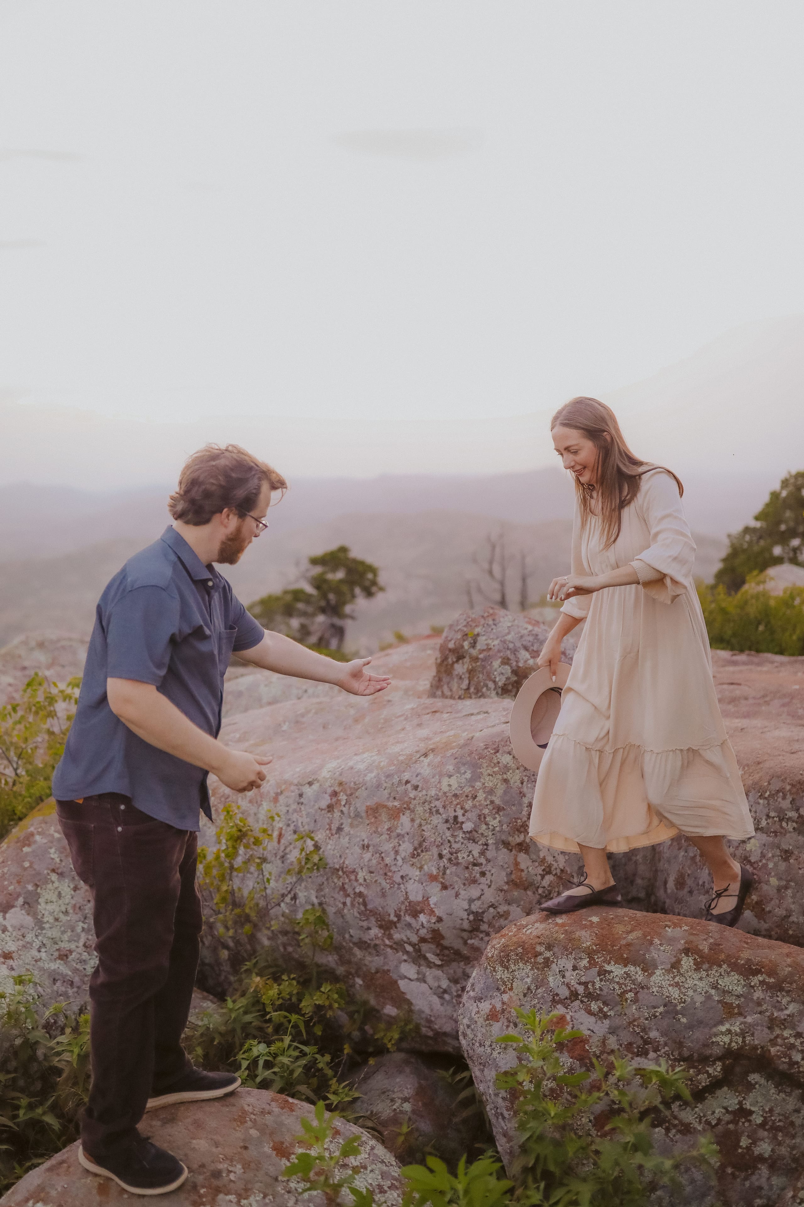 He offers a hand as she steps across the rocks, hat in hand