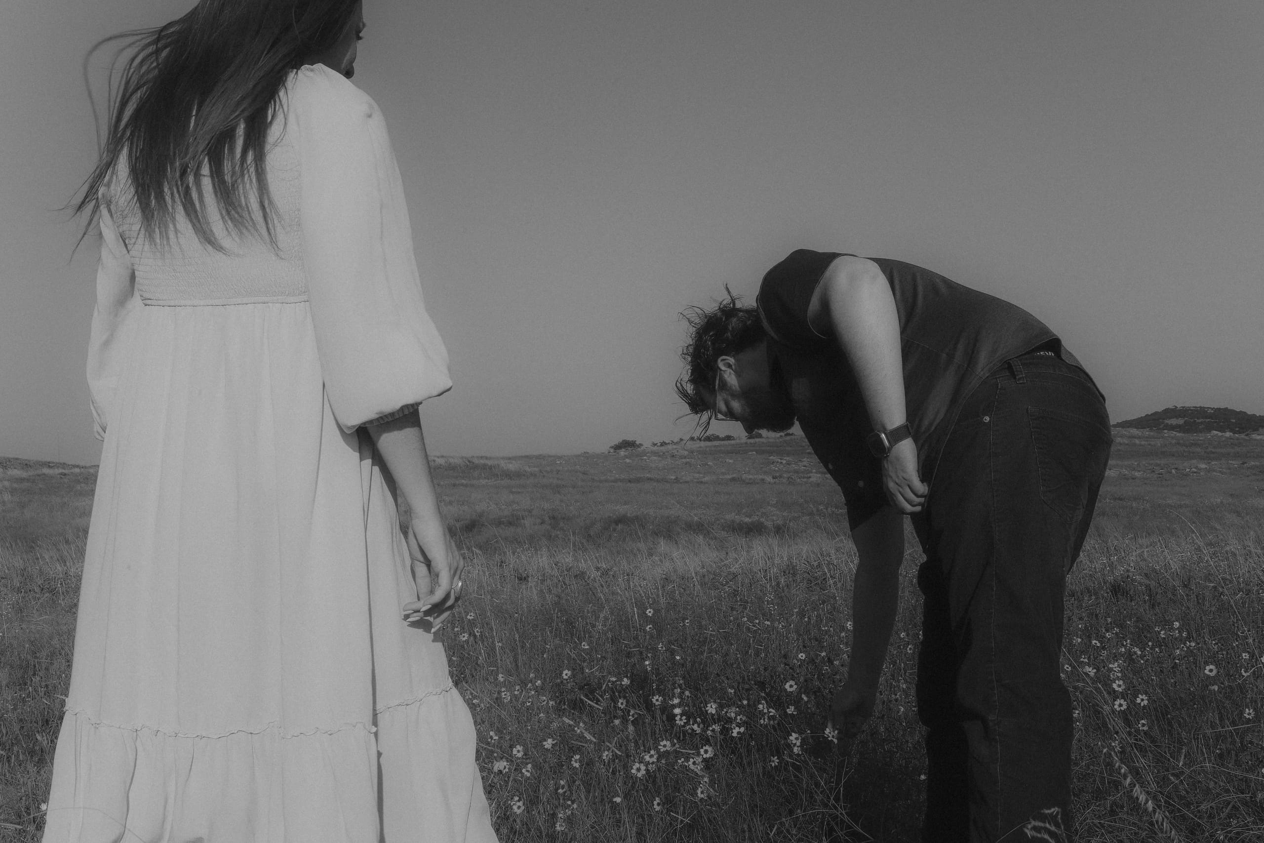 He stoops to pick meadow flowers while she looks on, in black and white