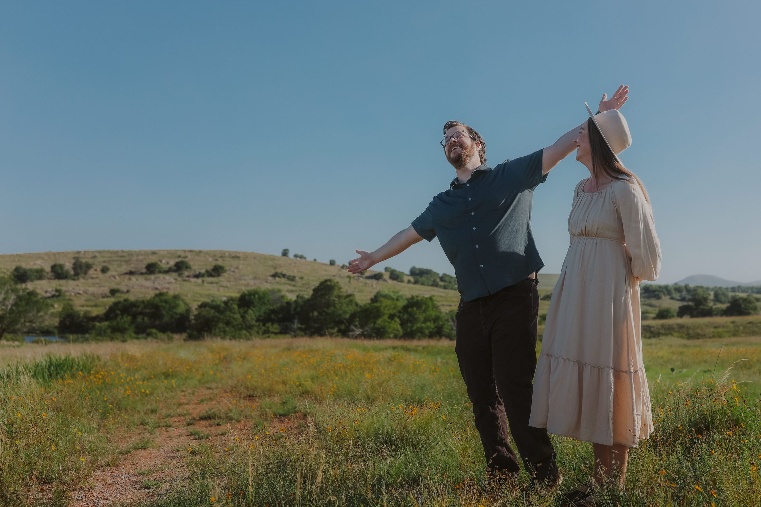 Arms flung wide on a grassy path, her hand holding on to her hat