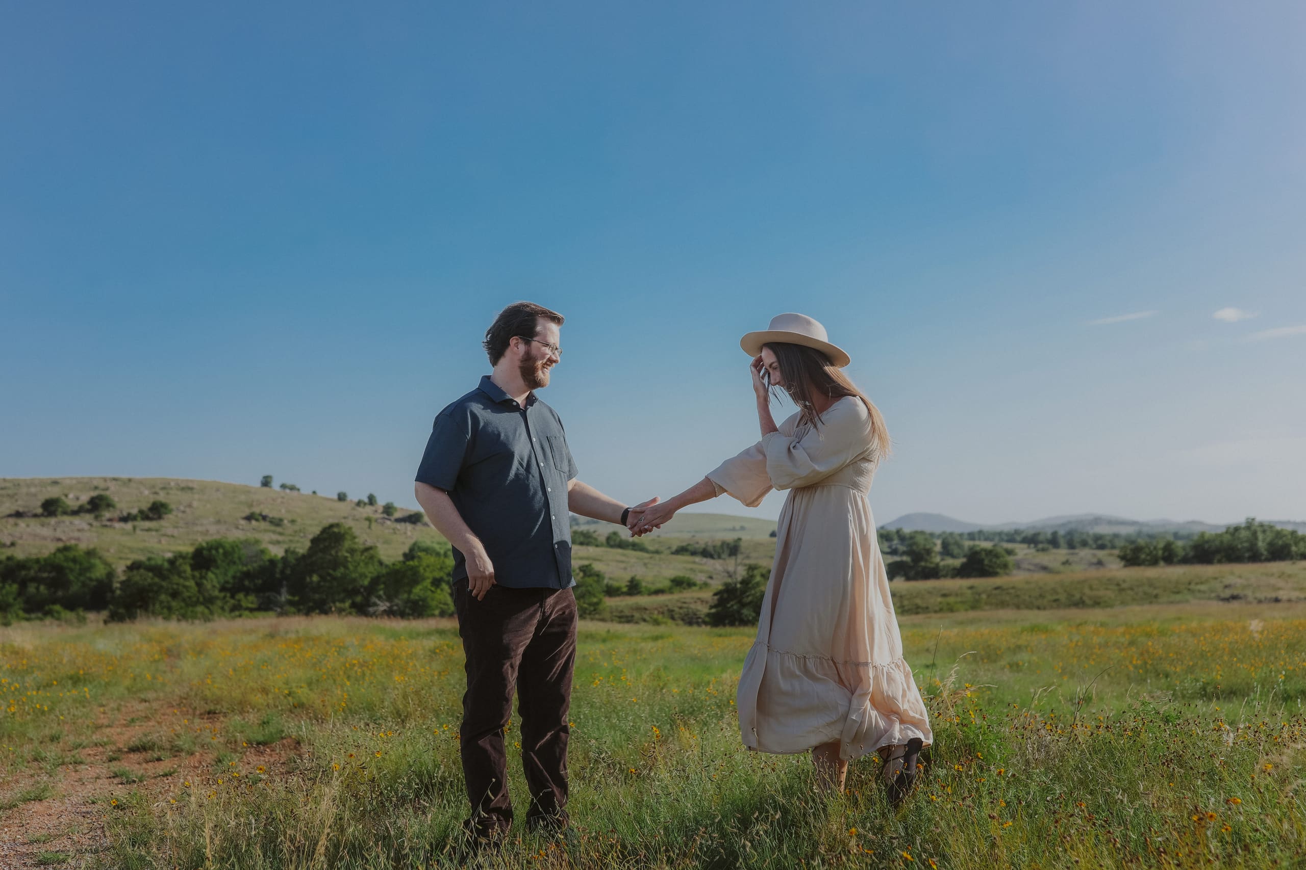 He takes her hand in the meadow as she steadies her hat